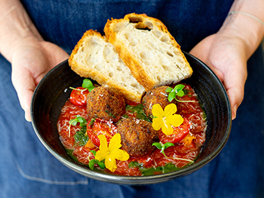 Eine Person in einer blauen Schürze hält eine schwarze Schüssel mit Brotscheiben, Tomatensoße, frittierten Bällchen und gelben essbaren Blüten in der Hand, garniert mit frischen Kräutern.
