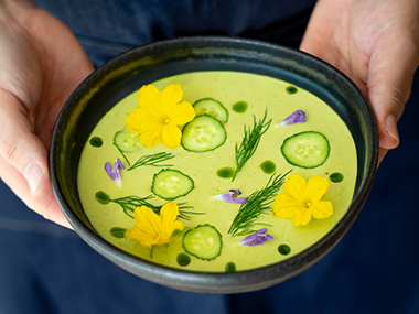 Eine Person hält eine Schüssel mit cremiger grüner Suppe in der Hand, die mit Gurkenscheiben, gelben essbaren Blüten, violetten Blütenblättern, frischem Dill und kleinen Tropfen grünen Öls garniert ist.