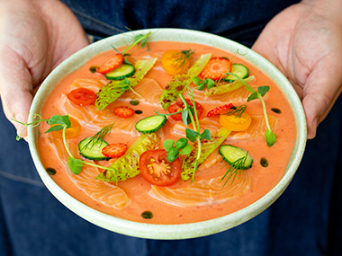 Eine Person hält eine Schüssel mit leuchtend rotem Lachsforellen Crudo in der Hand, garniert mit dünnen Lachs, geschnittenen Kirschtomaten, Gurke, Salat, Erdbeeren, Microgreens und einigen Tropfen Kräuteröl.