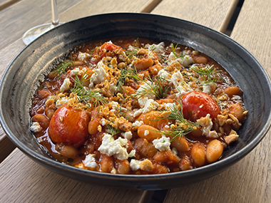 Eine schwarze Schüssel, gefüllt mit Baked Beans in Tomatensoße, garniert mit zerbröckeltem weißem Käse, Kräutern und einer Prise Gewürzen, steht auf einem Holztisch.