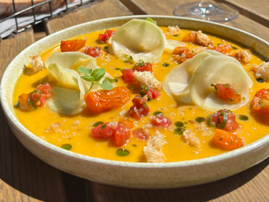 Eine flache Schüssel mit leuchtend gelber Suppe, garniert mit dünnen Gemüsestreifen, Tomatenwürfeln, frischen Kräutern und Croutons, auf einem Holztisch im Sonnenlicht.