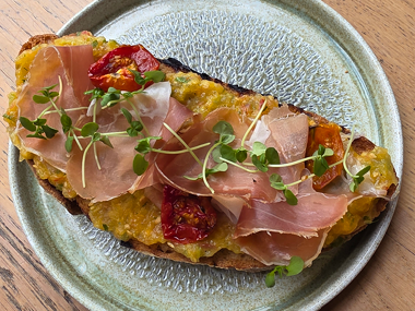 Eine Scheibe geröstetes Brot, belegt mit Prosciutto, gerösteten Tomaten, Microgreens und einem gelben Aufstrich, serviert auf einem Keramikteller.