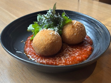In einer schwarzen Schüssel liegen zwei goldbraune Arancini auf einem Bett aus roter Tomatensauce, garniert mit geriebenem Käse und frischen grünen Kräutern.