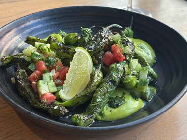 Auf einem Holztisch steht eine schwarze Schüssel mit gebratenen grünen Paprikaschoten, gewürfelten Tomaten, Gurke, grüner Soße und einer Limettenscheibe.