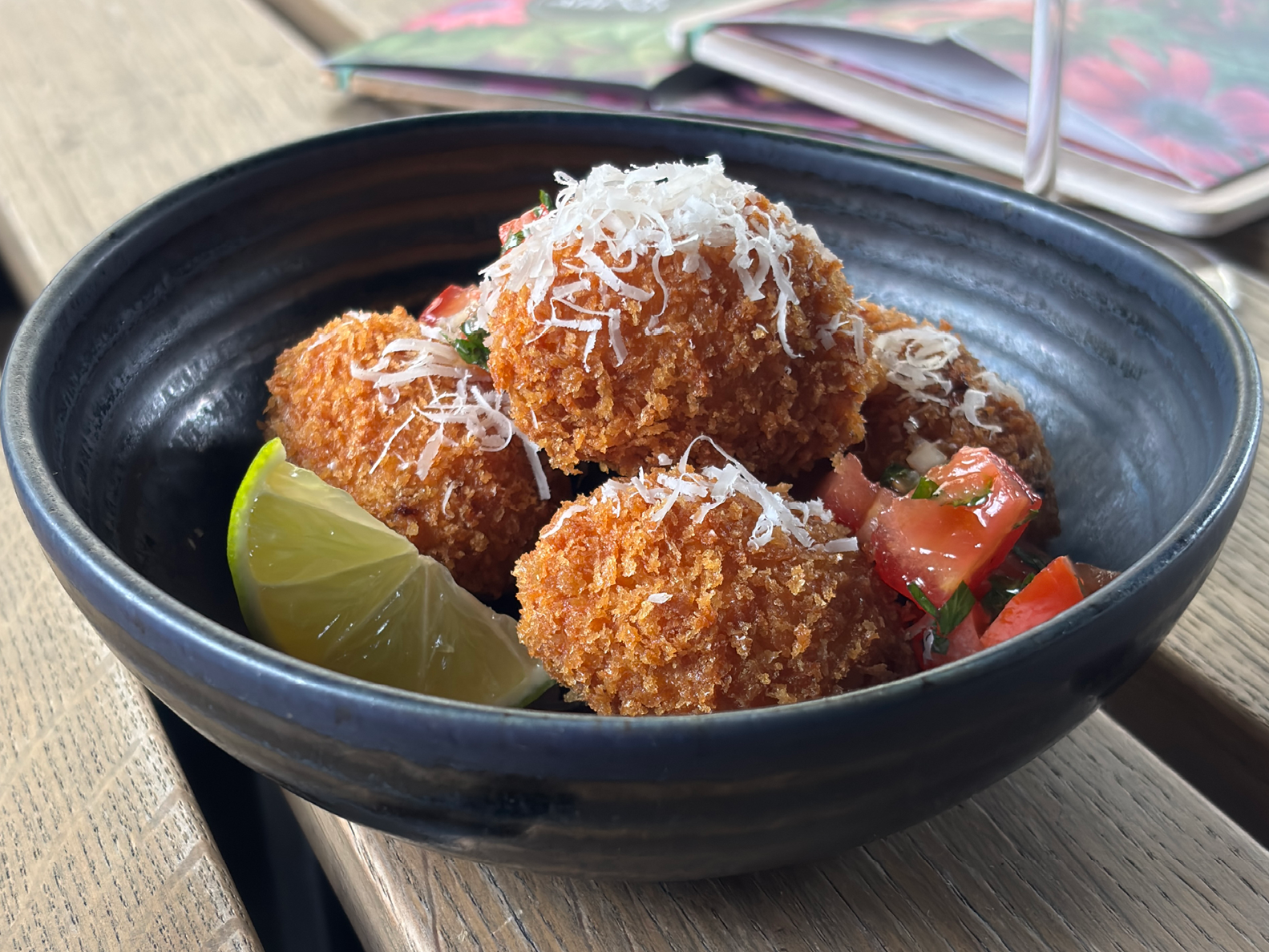 Eine schwarze Schüssel mit vier goldbraunen Kroketten, die mit geriebenem Käse bestreut sind, serviert mit Tomatenwürfeln, Kräutern und einer Limettenscheibe auf einem Holztisch.