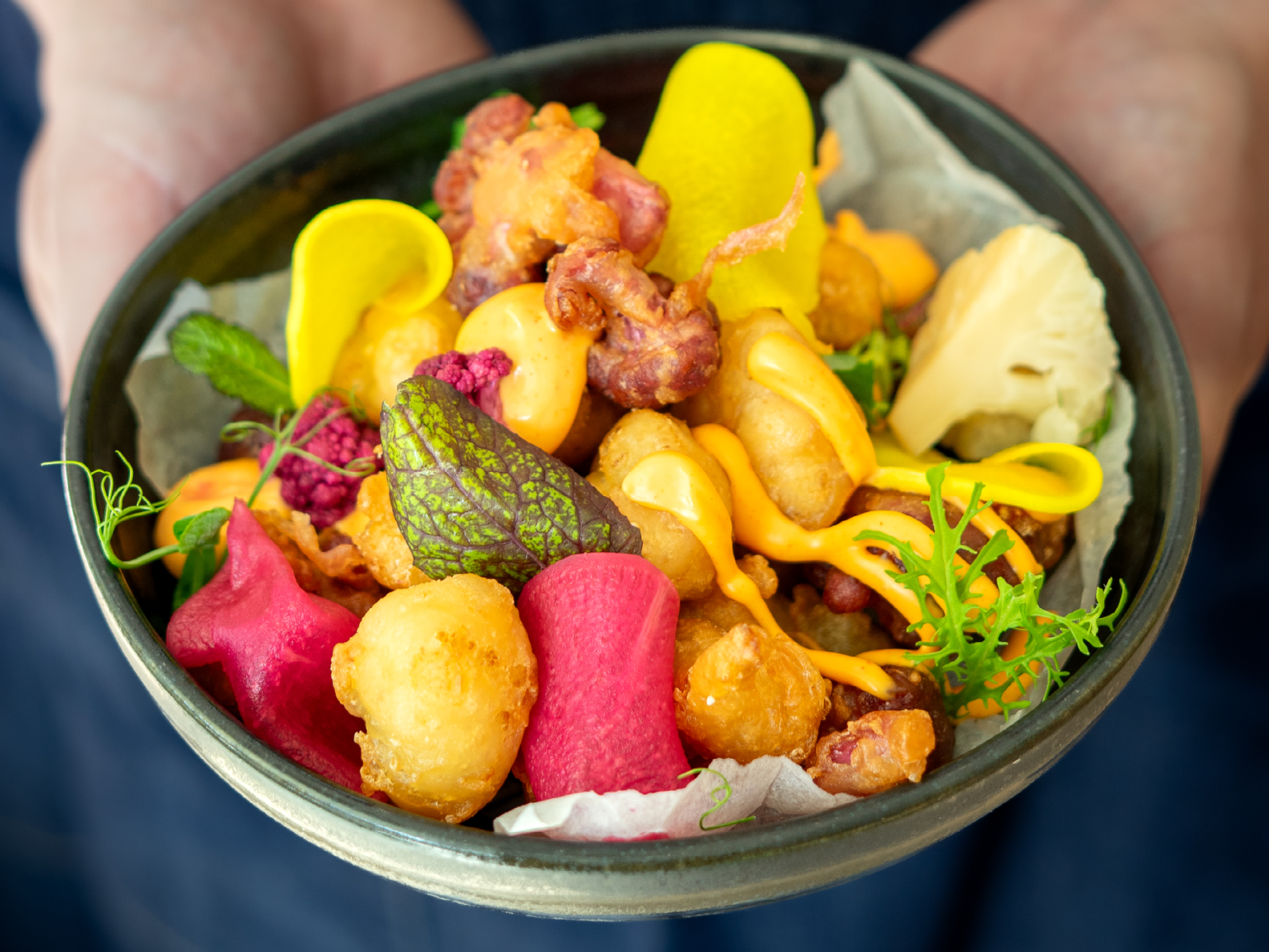 Eine mit beiden Händen gehaltene Schüssel, gefüllt mit gebratenem Blumenkohl, gelben und rosa Gemüsechips sowie Klecksen orangefarbener Soße.
