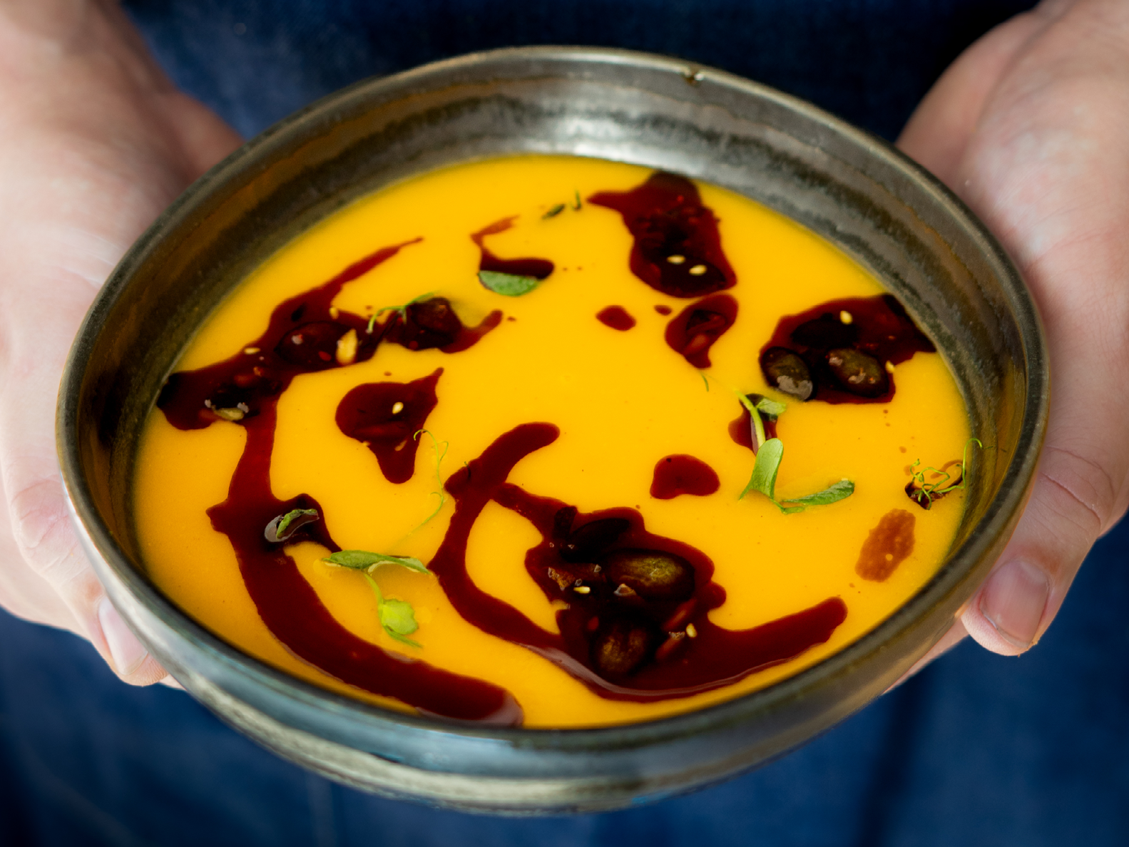 Hände, die eine Schüssel mit orangefarbener Suppe halten, über die dunkles Öl geträufelt und mit Samen und Kräutern garniert wurde.