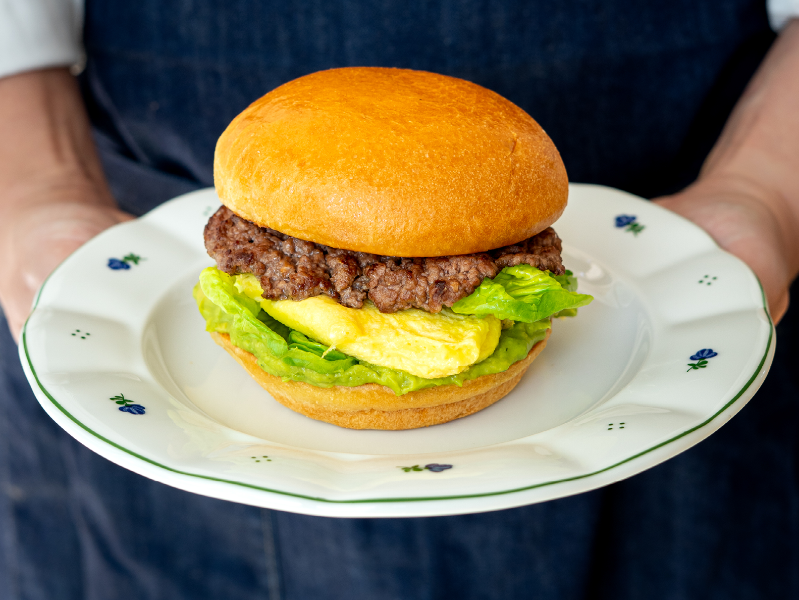 Eine Person, die einen weißen Teller mit einem Burger in der Hand hält, bestehend aus einem Rindfleisch-Patty, einem gefalteten Ei und Salat auf einem Brötchen.