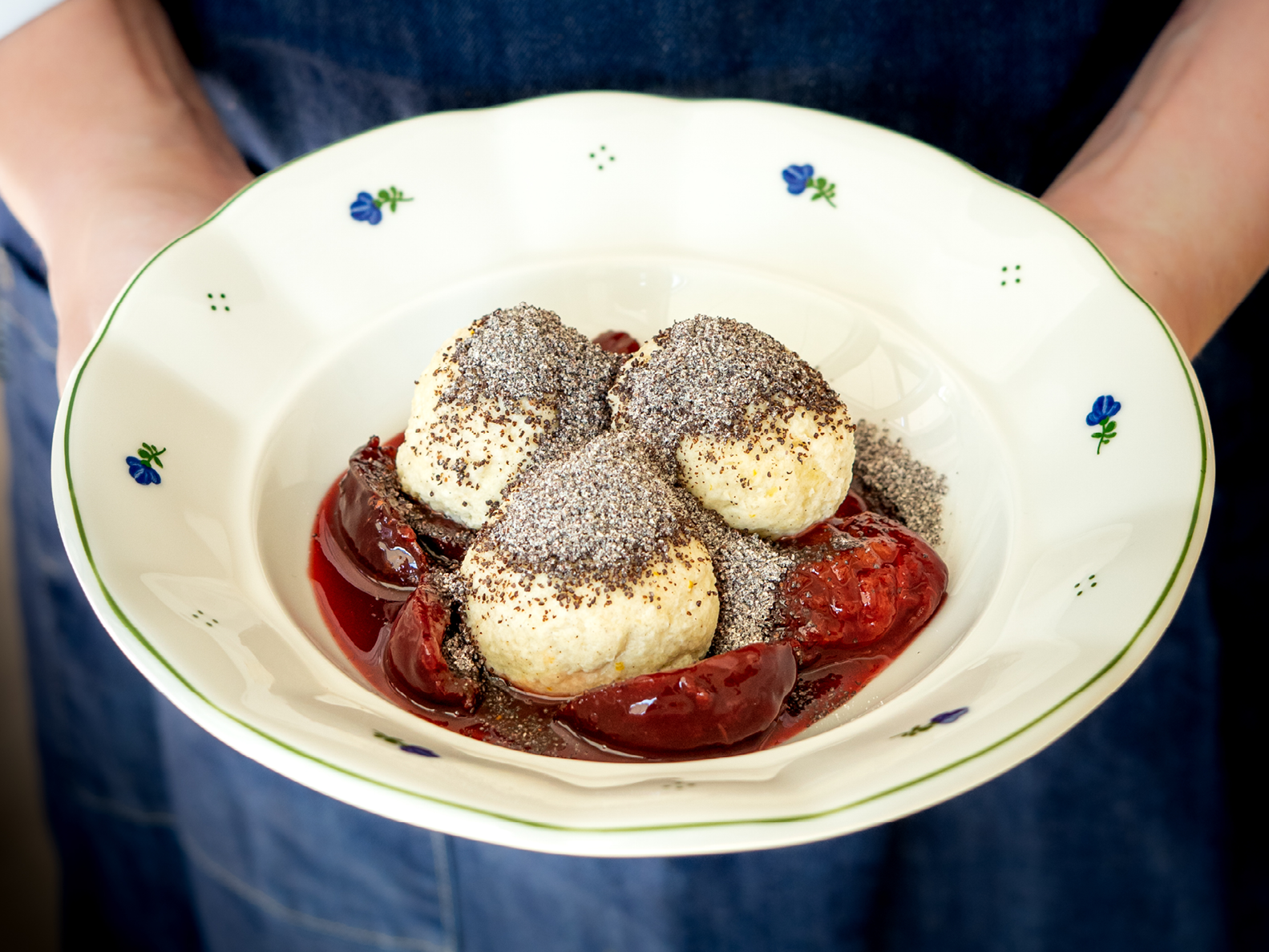Drei mit Mohn bestreute Knödel, serviert in einer Soße aus roten Beeren in einer Schale mit Blumenrand, die von einer Person in einer Jeansschürze gehalten wird.