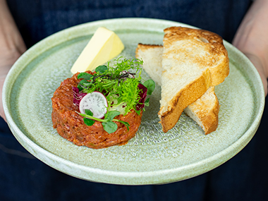 Ein grüner Teller mit Steak-Tartar, garniert mit Grünzeug und einer Radieschenscheibe, einem Stück Butter und zwei Scheiben geröstetem Brot.