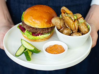 Eine Person hält einen weißen Teller mit einem Burger, gebratenen Fingerling-Kartoffeln in einer kleinen Schale, Gurkenscheiben und einer kleinen Schale mit Orangen-Dip-Sauce.
