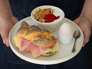 Eine Person hält einen Teller mit einem Sandwich aus Mohnbrot, Schinken und Käse, einem weichgekochten Ei in einem Eierbecher mit Löffel und einer Schale Joghurt mit Müsli und roter Fruchtsauce.