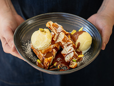 Eine Person hält eine dunkle Schale in der Hand, in der sich zwei Kugeln Vanilleeis, mit Schokoladensoße beträufelte und mit Puderzucker bestreute Waffelstücke und Pistazien befinden.