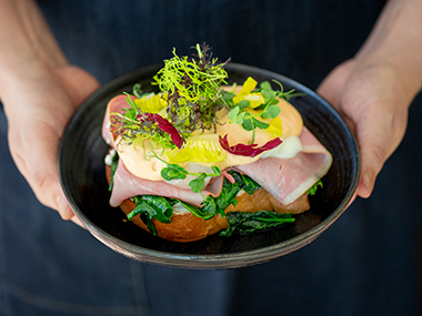 Eine Person hält einen schwarzen Teller mit einem offenen Sandwich, das mit Schinken, Blattgemüse, gelbem und rotem Gemüse und einer cremigen Soße belegt und mit frischen Kräutern garniert ist.