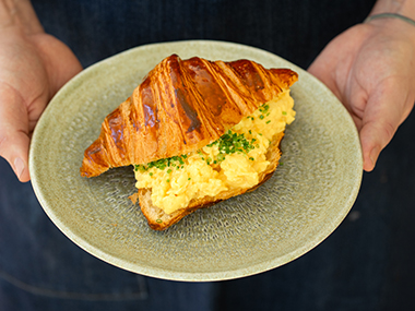 Eine Person hält einen Teller mit einem mit Rührei gefüllten Croissant, das mit gehacktem Schnittlauch bestreut ist.
