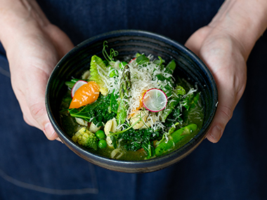 Eine Person hält eine schwarze Schale mit einem bunten Gemüsesalat, der mit geriebenem Käse bestreut und mit Radieschenscheiben und einer orangefarbenen Paprika garniert ist.