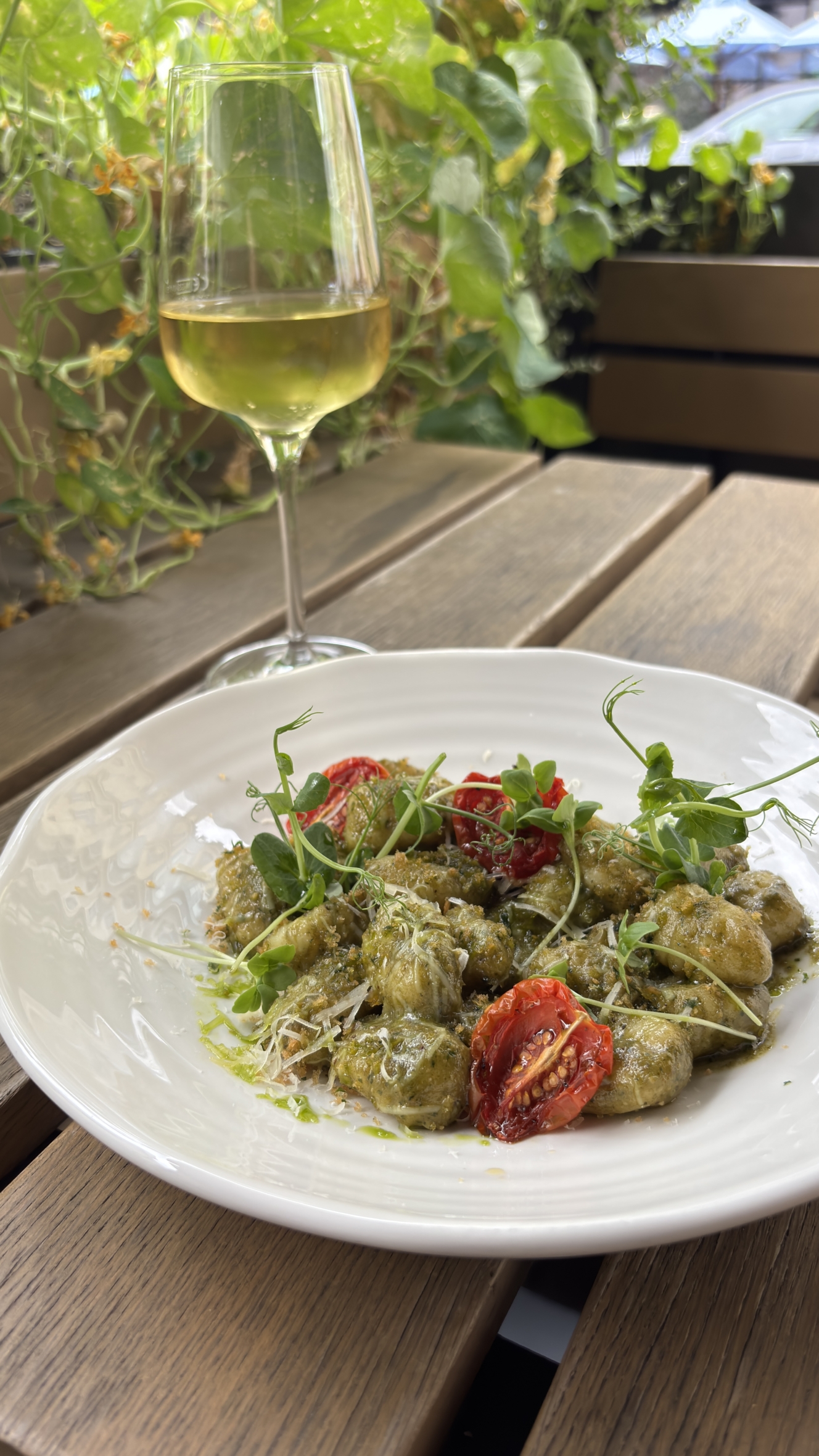 Auf einem Holztisch im Freien steht neben einem Glas Weißwein ein Teller mit Gnocchi, Pesto, gerösteten Tomaten und Blattgemüse. Im Hintergrund sind üppig grüne Pflanzen zu sehen.