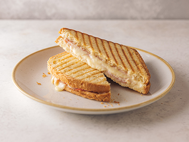 Ein Sandwich mit gegrilltem Käse und Schinken ist in zwei Hälften geschnitten und auf einen beigen Teller gestapelt. Das Brot hat goldene Grillspuren, und zwischen den Scheiben ist geschmolzener Käse zu sehen. Der Hintergrund ist eine helle, neutrale Fläche.