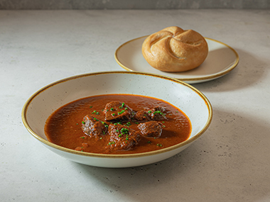 Eine Schüssel mit Rindergulasch, garniert mit gehackten Kräutern, steht auf einer hellen Fläche, im Hintergrund liegt ein rundes Brötchen auf einem Teller.