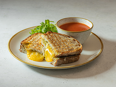 Ein Teller mit einem halbierten gegrillten Käsesandwich, aus dem der geschmolzene Käse herausläuft, serviert mit einem kleinen Salat und einem Teller Tomatensuppe.