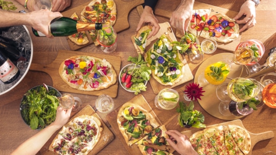 A group of people enjoys colorful flatbread pizzas topped with various vegetables and edible flowers, surrounded by drinks and fresh salads on a wooden table. One person pours sparkling wine into a glass.