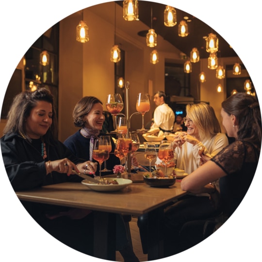 Vier Frauen sitzen an einem Restauranttisch, genießen Essen und Getränke, lächeln und unterhalten sich. Warme Beleuchtung und Hängelampen schaffen eine gemütliche, einladende Atmosphäre. Im Hintergrund ist ein Kellner zu sehen.