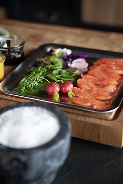 Auf einem Holzbrett steht ein Metalltablett mit Scheiben von rohem Lachs, Radieschen, frischem Grün und Kräutern. Im Vordergrund steht eine schwarze Schale, die mit grobem Salz gefüllt ist.