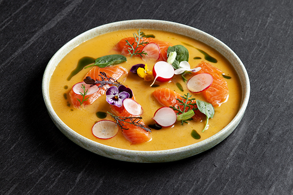 Eine flache Schale mit leuchtend orangefarbener Suppe, belegt mit Scheiben von rohem Lachs, Rettich, essbaren Blüten, Mikrogrün und kleinen Tropfen von grünem Öl, präsentiert auf einer dunklen Steinoberfläche.