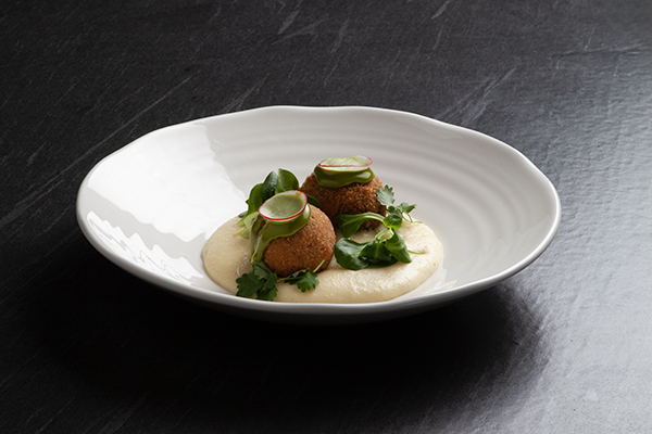 Drei goldbraune Kroketten mit geschnittenem Rettich, Mikrogrün und Kräutern, serviert auf einem cremigen weißen Püree in einer weißen, strukturierten Schale auf dunkler Oberfläche.