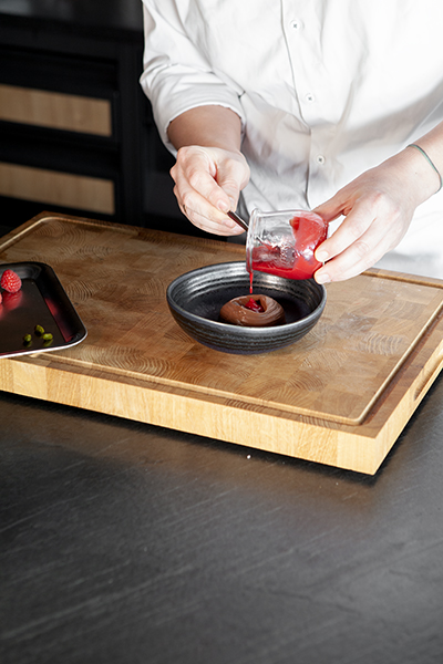Eine Person in einem weißen Kochkittel gießt rote Soße aus einem kleinen Glasgefäß auf ein Dessert in einer schwarzen Schüssel auf einem hölzernen Schneidebrett. Ein Metalltablett mit Himbeeren und grüner Garnitur steht daneben.