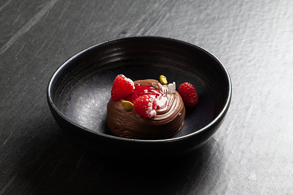In einer schwarzen Schale befindet sich eine Mousse au Chocolat, die mit drei frischen Himbeeren, roter Sauce, grünen Pistazien und Meersalzflocken auf einer dunklen, strukturierten Oberfläche angerichtet ist.