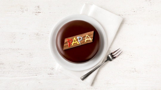 Ein runder Schokoladenkuchen auf einem weißen Teller, gekrönt von einer Holztafel mit der Aufschrift PAPA in bunter Glasur. Der Teller steht auf einer weißen Serviette mit einer Gabel daneben, alles auf einer weißen Holzfläche.
