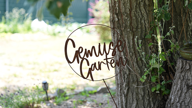 In einem Garten steht neben einem Baum ein Metallschild mit der Aufschrift „Gemüse Garten“, im Hintergrund sind grünes Laub und verschwommene Elemente der Außenlandschaft zu sehen.