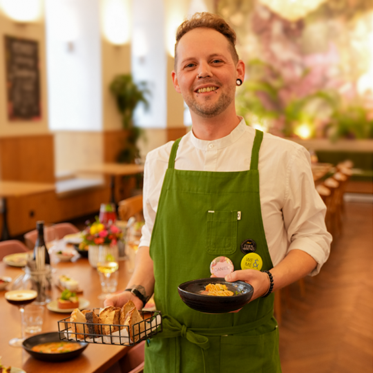 Ein lächelnder Kellner in einer grünen Schürze hält einen Korb mit Brot und eine Schüssel mit Essen in einem warm beleuchteten Restaurant, in dessen Hintergrund gedeckte Tische und Blumenschmuck zu sehen sind.