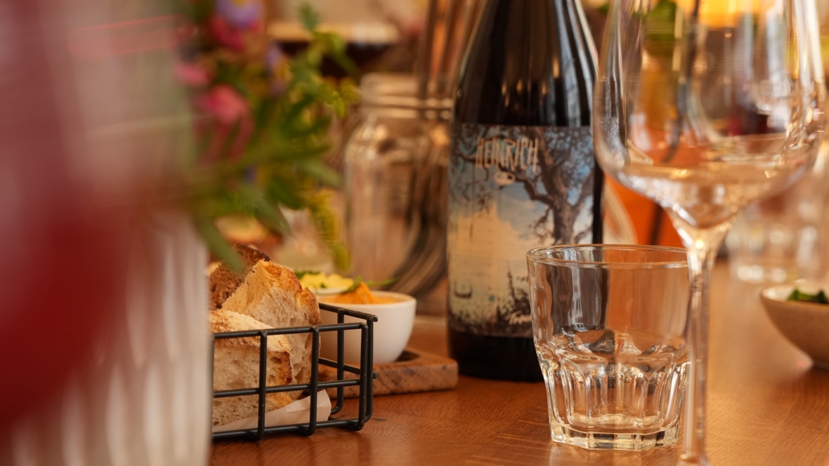 Eine Weinflasche mit der Aufschrift Heinrich neben einem leeren Glas, einem kleinen Glas Wasser und einem Drahtkorb mit Brotscheiben.